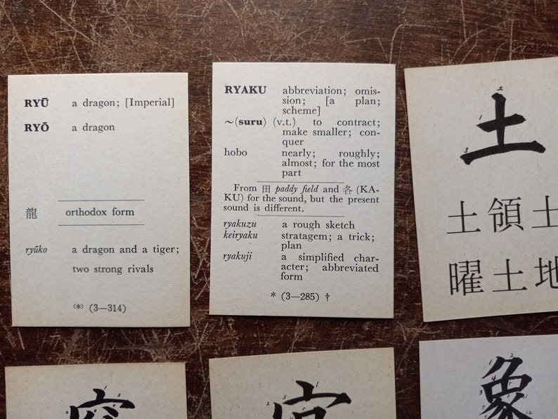 May include: Several small white cards with Japanese text and English translations are arranged on a textured brown surface. The cards feature words like "dragon" and "abbreviation", with corresponding definitions.