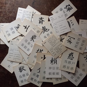 May include: A collection of vintage flashcards with Japanese characters and English definitions. The cards are scattered on a dark wooden surface, with the black ink characters standing out against the aged paper.