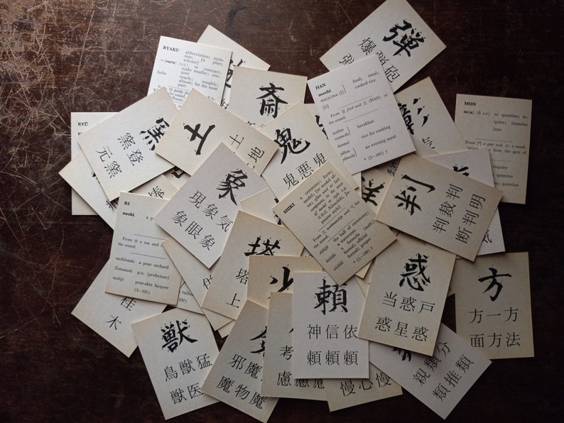 May include: A collection of vintage flashcards with Japanese characters and English definitions. The cards are scattered on a dark wooden surface, with the black ink characters standing out against the aged paper.
