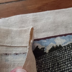 May include: A close-up of a white linen fabric with a folded edge. The fabric has a printed design of a building with a dark brown roof and a black and white patterned facade.