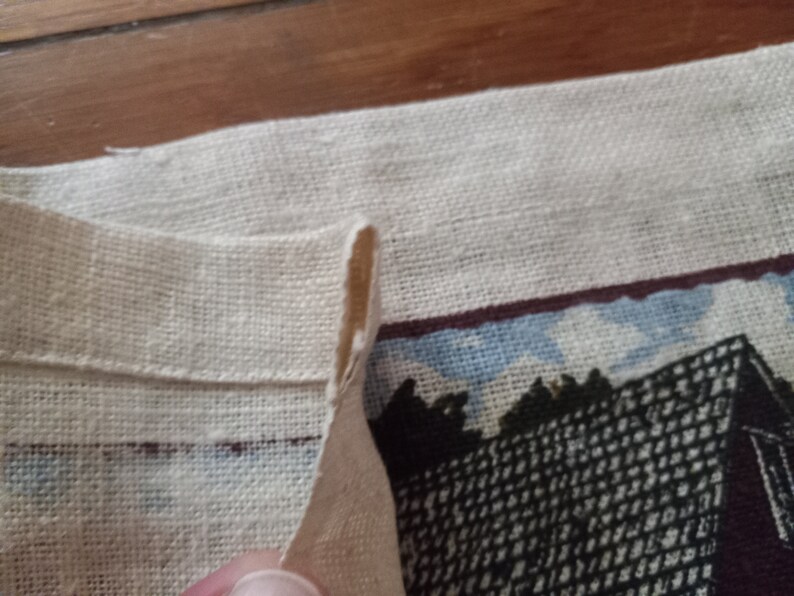 May include: A close-up of a white linen fabric with a folded edge. The fabric has a printed design of a building with a dark brown roof and a black and white patterned facade.