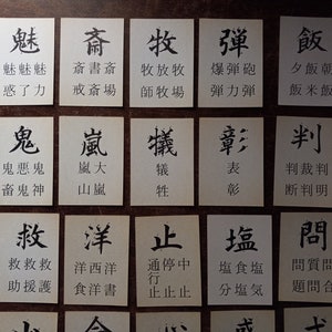 May include: A collection of small, rectangular cards with black Japanese characters printed on them. The cards are arranged on a dark wooden surface, with each card displaying a different character and its associated words.