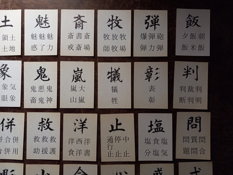 May include: A collection of small, rectangular cards with black Japanese characters printed on them. The cards are arranged on a dark wooden surface, with each card displaying a different character and its associated words.