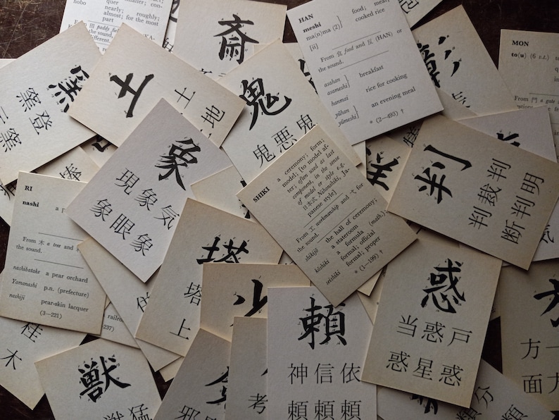 May include: A collection of vintage flashcards with Japanese characters and English definitions. The cards are arranged in a scattered pattern, with the black ink characters standing out against the aged paper.