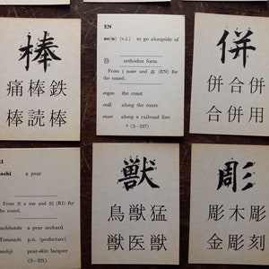 May include: A collection of flashcards with Japanese characters and English definitions. The cards are arranged on a dark wooden surface. Each card features a different character with its pronunciation and meaning.