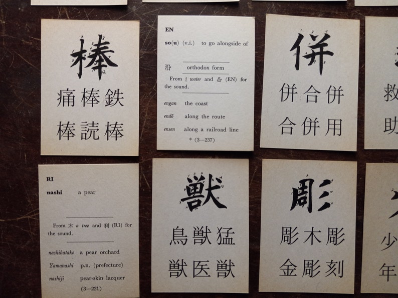 May include: A collection of flashcards with Japanese characters and English definitions. The cards are arranged on a dark wooden surface. Each card features a different character with its pronunciation and meaning.