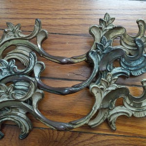 May include: Two ornate, antique bronze drawer pulls with a distressed finish. The pulls feature a scrolling, floral design with curved handles. The hardware is set against a wooden surface.
