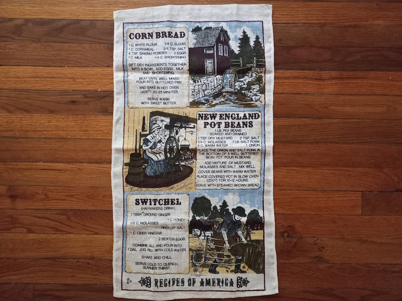 May include: A vintage-style tea towel with a beige background and brown text. The towel features three recipes: Cornbread, New England Pot Beans, and Switchel. Each recipe is accompanied by a small illustration.