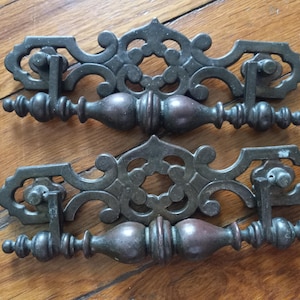 May include: Antique bronze-toned drawer pulls with ornate, scrollwork designs. Each pull features a central handle with decorative ends and a detailed backplate. These vintage hardware pieces are ideal for furniture restoration projects.