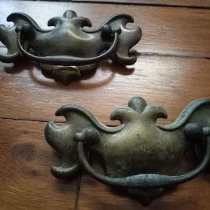 May include: Two antique bronze drawer pulls with ornate, curved designs. Each pull features a central handle and decorative flourishes, suggesting a vintage or classic style. The pulls are set against a wooden surface.