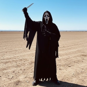 Puede incluir: Una persona con una túnica negra con capucha y una máscara blanca, sosteniendo un cuchillo en el aire. El disfraz se encuentra sobre un fondo de un campo marrón y árido y un cielo azul claro, evocando un tema de terror.