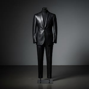 May include: A black leather suit displayed on a mannequin. The suit includes a two-button blazer with notched lapels and matching trousers. The suit is a solid black color and appears to be made of high-quality leather. The mannequin is set against a dark gray background.