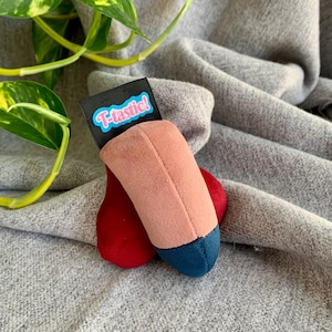 Puede incluir: Un juguete de peluche con forma de modelo anatómico estilizado. El juguete presenta un cuerpo rosa, una base roja y una punta azul. Una etiqueta negra con el texto "T-tastic!" está adjunta.