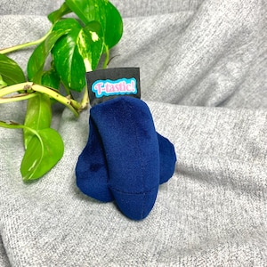 Puede incluir: Un objeto de felpa azul oscuro con una textura suave similar al terciopelo. En la parte superior tiene una etiqueta de tela negra con el texto T-tastic! en fuente azul claro con borde rosa. El objeto tiene una forma redondeada de tres lóbulos.
