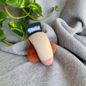 Puede incluir: Un juguete de peluche con forma de pene, con un eje beige, una punta rosa y testículos marrones. Una etiqueta negra con la palabra "T-tastic!" en azul y blanco está adjunta. El juguete está sobre un fondo de tela gris.