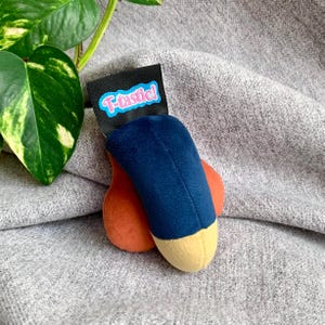 Puede incluir: Un juguete de peluche con forma de fruta o verdura estilizada, con un cuerpo azul marino, una punta beige y una base naranja. Una etiqueta negra con el texto "T-tastic!" en azul y rosa está adjunta.
