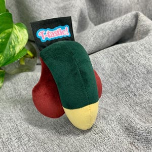 Può includere: Un peluche caratterizzato da una sezione centrale verde scuro, una punta gialla e due componenti laterali rossi arrotondati. Un'etichetta rettangolare nera è fissata in alto, riportante la scritta T-tastic! in lettere rosa con bordo blu.