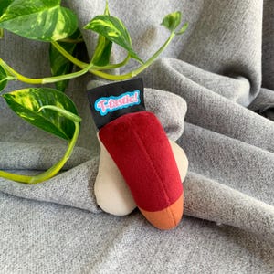 Puede incluir: Un juguete de peluche con forma de paleta roja con base beige y punta naranja. Una etiqueta negra con la palabra "T-tastic!" en azul y blanco está adjunta. El juguete está sobre un fondo de tela gris.