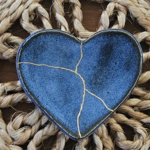 Könnte beinhalten: Eine herzförmige Keramikschale mit einer texturierten blauen Glasur. Die Oberfläche weist sichtbare Risse auf, die mit goldfarbenen Linien repariert wurden und ein markantes Muster in der Mitte des Stücks bilden. Die Ränder sind dunkel und erhöht.