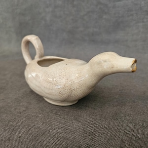 Könnte beinhalten: Eine keramikente-förmige Sahnekännchen mit Henkel und Ausguss. Das Sahnekännchen ist cremefarben mit einer Craquelé-Glasur und einem goldfarbenen Rand am Ausguss. Die Ente ruht auf einer grauen Oberfläche.