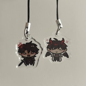 Puede incluir: Dos colgantes transparentes de acrílico con cordones negros. Cada colgante presenta un personaje de dibujos animados con cabello oscuro y detalles rojos. Un personaje tiene una espada y una expresión de enfado, el otro tiene alas y una cara sonriente.