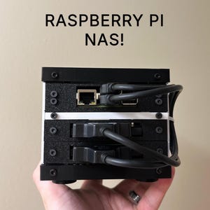 Könnte beinhalten: Ein schwarz-weißes Raspberry Pi NAS-Gerät mit mehreren Anschlüssen und angeschlossenen Kabeln. Das Gerät ist gestapelt und mit Schrauben zusammengehalten. Der Text "RASPBERRY PI NAS!" ist oben aufgedruckt.