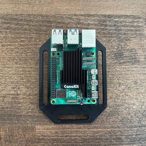 May include: A green Raspberry Pi circuit board with a black heat sink and the text "CanaKit" is mounted in a black plastic case. The board has multiple ports and connectors. The case has mounting holes.