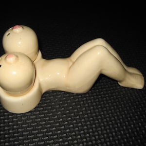 Könnte beinhalten: Ein Salz- und Pfefferstreuer-Set aus Keramik in Form einer liegenden Figur mit Sockel. Die Figur ist cremefarben mit rosa Akzenten.