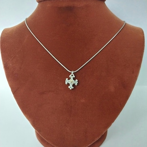 Puede incluir: Un collar de plata con un peque&ntilde;o colgante de cruz ornamentado. El colgante presenta un centro dorado y detalles intrincados. El collar se exhibe sobre un busto de terciopelo marr&oacute;n.