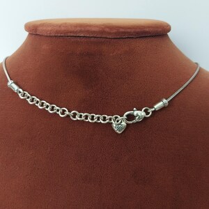 Puede incluir: Collar plateado con un peque&ntilde;o dije en forma de coraz&oacute;n. El collar presenta una cadena delicada y una serie de peque&ntilde;os anillos unidos cerca del cierre. El dije de coraz&oacute;n tiene un dise&ntilde;o grabado. El collar se exhibe en un maniqu&iacute; marr&oacute;n.