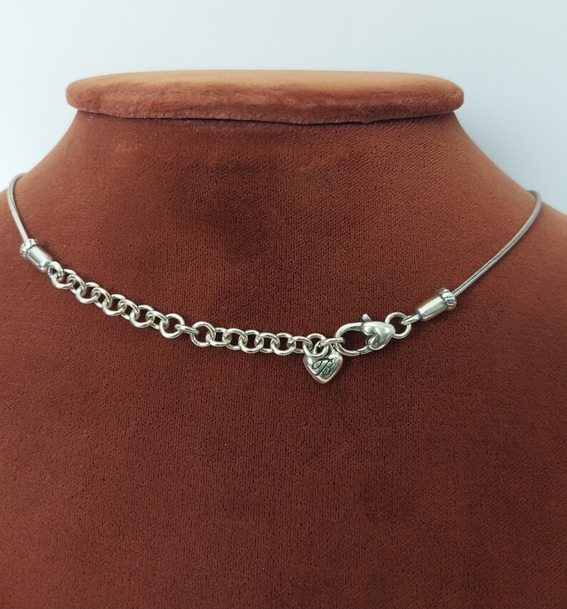 Puede incluir: Collar plateado con un peque&ntilde;o dije en forma de coraz&oacute;n. El collar presenta una cadena delicada y una serie de peque&ntilde;os anillos unidos cerca del cierre. El dije de coraz&oacute;n tiene un dise&ntilde;o grabado. El collar se exhibe en un maniqu&iacute; marr&oacute;n.
