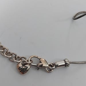 Puede incluir: Collar plateado con un dije en forma de coraz&oacute;n y un cierre de mosquet&oacute;n. El dije tiene un dise&ntilde;o decorativo y el collar tiene una cadena y una cadena de serpiente. El collar est&aacute; sobre una superficie blanca.