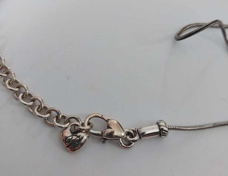 Puede incluir: Collar plateado con un dije en forma de coraz&oacute;n y un cierre de mosquet&oacute;n. El dije tiene un dise&ntilde;o decorativo y el collar tiene una cadena y una cadena de serpiente. El collar est&aacute; sobre una superficie blanca.