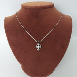 Puede incluir: Un collar plateado con un colgante de cruz. El colgante presenta una gema transparente central y detalles ornamentados. El collar se exhibe sobre un busto marr&oacute;n sobre un fondo blanco, mostrando el dise&ntilde;o y la artesan&iacute;a de la joya.