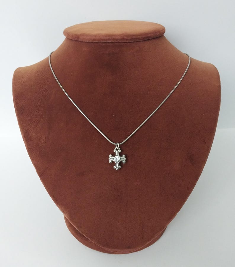 Puede incluir: Un collar plateado con un colgante de cruz. El colgante presenta una gema transparente central y detalles ornamentados. El collar se exhibe sobre un busto marr&oacute;n sobre un fondo blanco, mostrando el dise&ntilde;o y la artesan&iacute;a de la joya.
