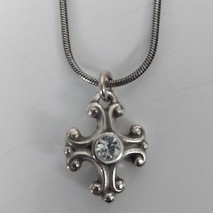 Puede incluir: Collar plateado con un colgante de cruz decorativa con una gema transparente en el centro. La cruz tiene un dise&ntilde;o ornamental con volutas. La cadena es una fina cadena de serpiente texturizada. El colgante mide aproximadamente 2,5 cm de largo.