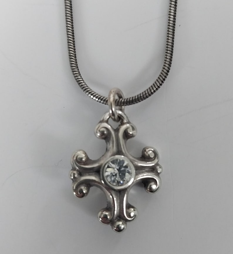 Puede incluir: Collar plateado con un colgante de cruz decorativa con una gema transparente en el centro. La cruz tiene un dise&ntilde;o ornamental con volutas. La cadena es una fina cadena de serpiente texturizada. El colgante mide aproximadamente 2,5 cm de largo.