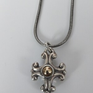 Puede incluir: Collar con colgante plateado con un dise&ntilde;o de cruz decorativa y un centro dorado. El colgante cuelga de una cadena de serpiente plateada. La cruz tiene detalles ornamentados y arremolinados.