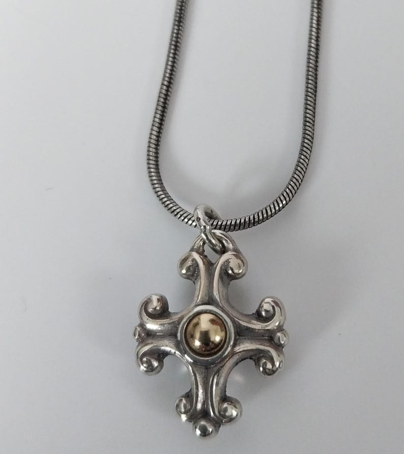 Puede incluir: Collar con colgante plateado con un dise&ntilde;o de cruz decorativa y un centro dorado. El colgante cuelga de una cadena de serpiente plateada. La cruz tiene detalles ornamentados y arremolinados.