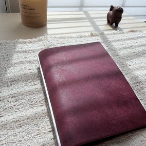 May include: A rectangular, deep burgundy notebook with a smooth, matte cover and a silver-colored spine. The notebook is positioned horizontally, showcasing its solid, dark red front surface and the metallic edge detail along the left side.