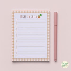 May include: A lined notepad with a tan gingham border. The notepad features the text "NEGES I'W GOFIO" and a small illustration of a basket of vegetables. A pink pen lies next to the notepad.