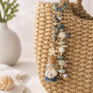 May include: A decorative keyring featuring a small glass bottle filled with tiny seashells and blue stones, topped with a cork stopper and a white starfish charm. It includes a strand of sage green, clear crackle, and gold beads with shell accents.
