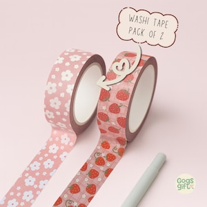 May include: Two rolls of washi tape with decorative patterns. One roll features white flowers on a pink background, and the other has red strawberries on a pink grid. The text "Washi Tape Pack of 2" is displayed above the strawberry tape.