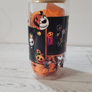 Puede incluir: Un vaso de vidrio transparente con tapa de bamb&uacute; y pajita. El vaso presenta un dise&ntilde;o envolvente con personajes e im&aacute;genes de Pesadilla antes de Navidad, incluyendo a Jack Skellington y calabazas. El vaso contiene papel de seda naranja.