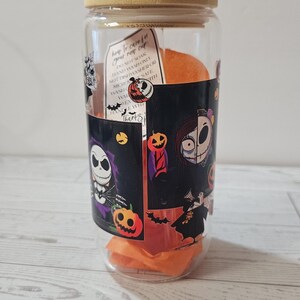 Puede incluir: Un vaso de vidrio transparente con tapa de bamb&uacute; y pajita de vidrio. El vaso est&aacute; decorado con personajes e im&aacute;genes tem&aacute;ticas de Halloween. Dentro del vaso hay pa&ntilde;uelos de papel naranja. Una etiqueta en el vaso dice "how to care for your new cup".