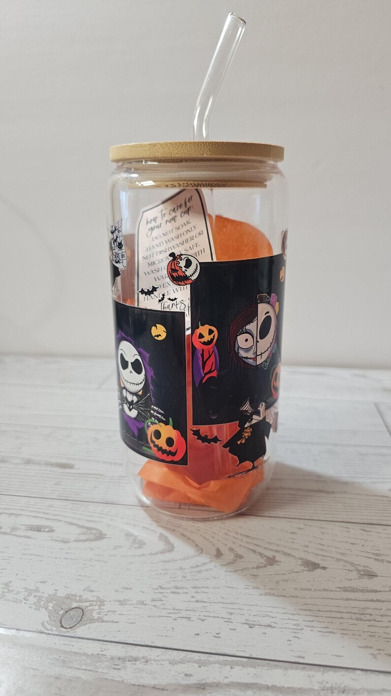 Puede incluir: Un vaso de vidrio transparente con tapa de bamb&uacute; y pajita de vidrio. El vaso est&aacute; decorado con personajes e im&aacute;genes tem&aacute;ticas de Halloween. Dentro del vaso hay pa&ntilde;uelos de papel naranja. Una etiqueta en el vaso dice "how to care for your new cup".