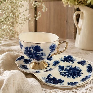 Puede incluir: Un juego de taza de té y plato de porcelana color crema con un patrón floral azul oscuro. La taza tiene un borde festoneado, una base de pedestal dorada y un asa dorada. El plato presenta un diseño floral azul a juego en toda su superficie.