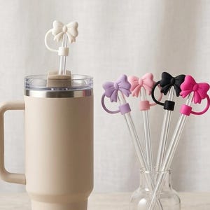 Peut inclure: Un gobelet isotherme beige avec une poignée et un couvercle transparent, à côté d'un vase en verre contenant plusieurs pailles transparentes surmontées de breloques en silicone en forme de nœud coloré. Les breloques sont dans les tons de rose, de violet et de noir.