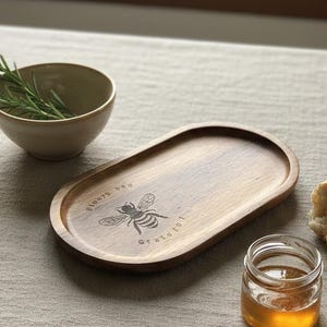 Può includere: Un vassoio di legno con un disegno di ape inciso e le parole «always be grateful». Un piccolo barattolo di miele, una ciotola con rametti di rosmarino e un pezzo di pane sono anche sul tavolo. Il vassoio è ovale.