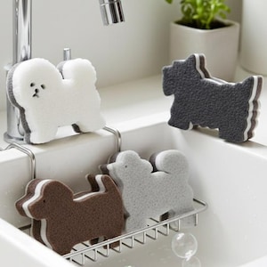 Peut inclure: Éponges de cuisine en forme de chien dans un évier blanc. Les éponges sont de différentes couleurs, dont le blanc, le gris et le marron. Une éponge est fixée à un support métallique, tandis que d'autres sont dans l'évier. Un robinet chromé est visible en arrière-plan.